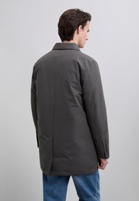 Man in een donkergrijze lange jas en blauwe spijkerbroek, staand met zijn rug gedeeltelijk gekeerd naar een effen lichtgrijze achtergrond.