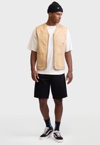 Chaleco de trabajo color tostado con dos bolsillos frontales, usado sobre una camiseta color crema, combinado con pantalones cortos negros y zapatillas negras y blancas.