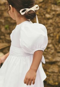 Robe blanche avec des manches bouffantes, une taille cintrée et un nœud dans le dos. Tissu doux au toucher lisse, présentée de côté.