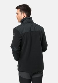 Zwarte fleecejas met een hoge kraag, contrasterend schouderpaneel en ritszakken aan de zijkant. Bevat een gestructureerde stof en een logopatch.
