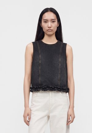 Femme aux longs cheveux noirs portant un haut en denim noir sans manches avec un ourlet à volants et un pantalon blanc, debout devant un fond uni.