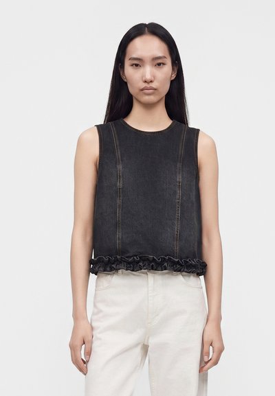 Femme aux longs cheveux noirs portant un haut en denim noir sans manches avec un ourlet à volants et un pantalon blanc, debout devant un fond uni.