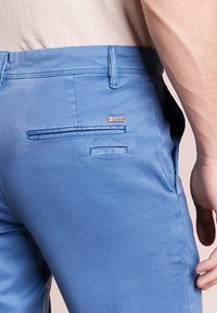 Modré bavlněné kalhoty s kapsou na zadní straně, která má skrytý design, a malou kovovou štítek u pasu. Hladká textilie.