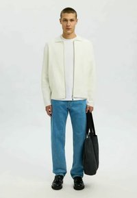 Valge tõmblukk-kampsun valge T-särgi, siniste teksade ja mustade kingade peal. Hoidis musta kott-äri kotti. Lihtne, kaasaegne disain puhaste joontega.