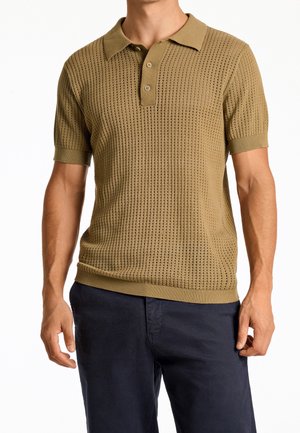 Beige stickad polotröja med ett texturerat mönster, korta ärmar och en placket med tre knappar; kombinerad med mörkblå byxor.