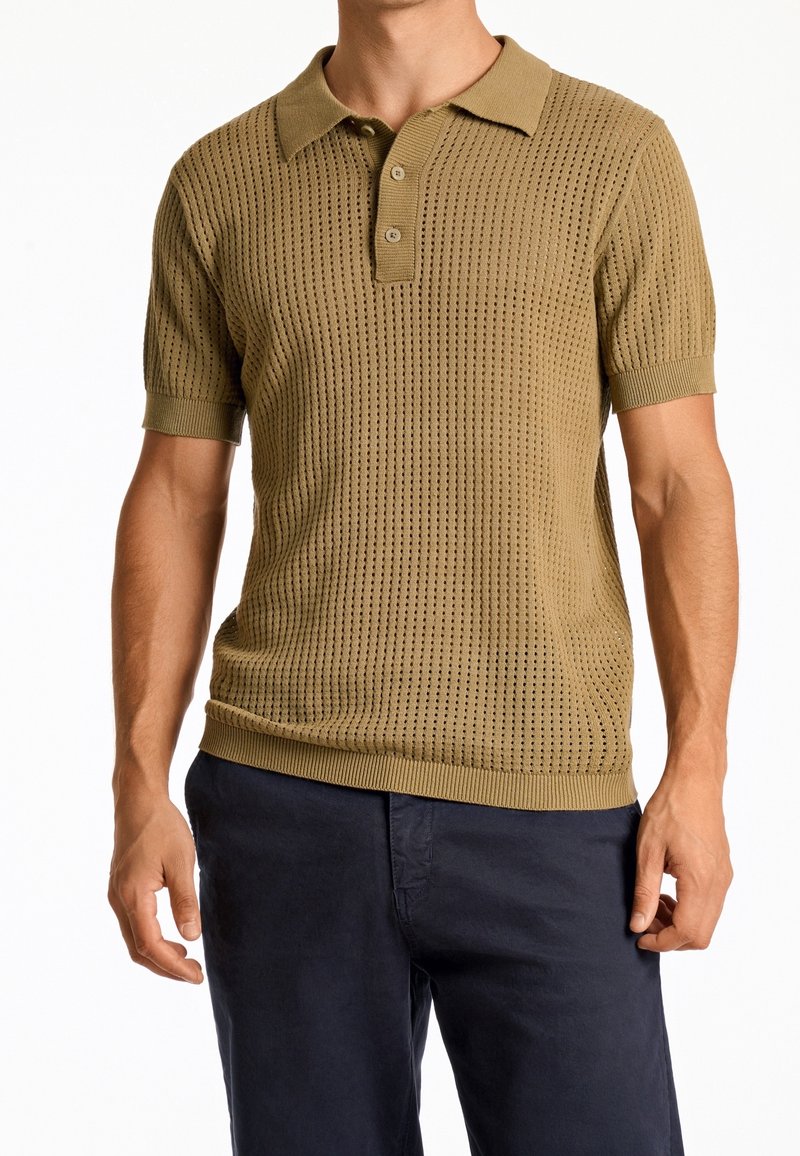 Beiger gestrickter Poloshirt mit strukturiertem Muster, kurzen Ärmeln und einer Drei-Knopf-Leiste; kombiniert mit dunkelblauen Hosen.