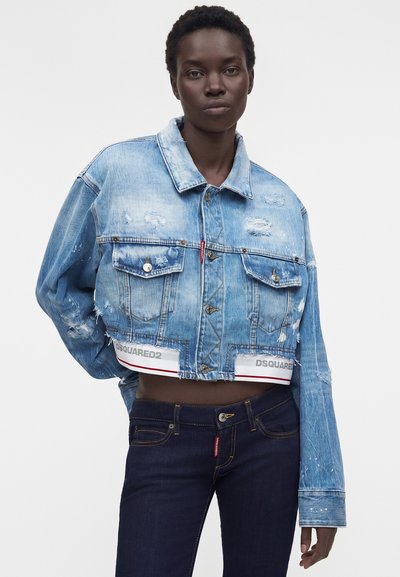 Γυναίκα που φοράει ένα φθαρμένο cropped τζιν μπουφάν σε ανοιχτό μπλε και σκούρο μπλε τζιν, στέκεται με το ένα χέρι πίσω από την πλάτη της μπροστά σε απλό φόντο.