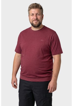 Mężczyzna z brodą w bordowej koszulce z krótkim rękawem i czarnych spodniach typu cargo, uśmiechający się i stojący na jasnym, jednolitym tle.