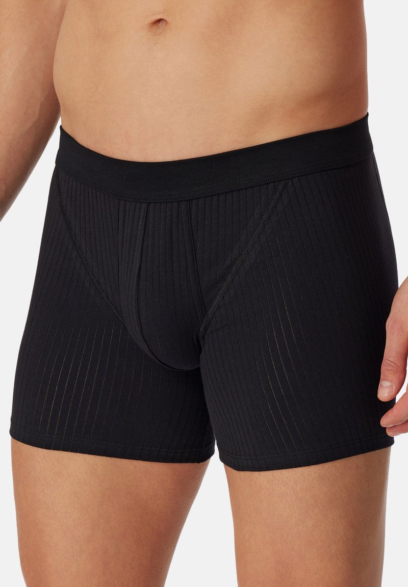 Torso maschile che indossa boxer aderenti neri con una vita elastica rifinita su uno sfondo chiaro e semplice.