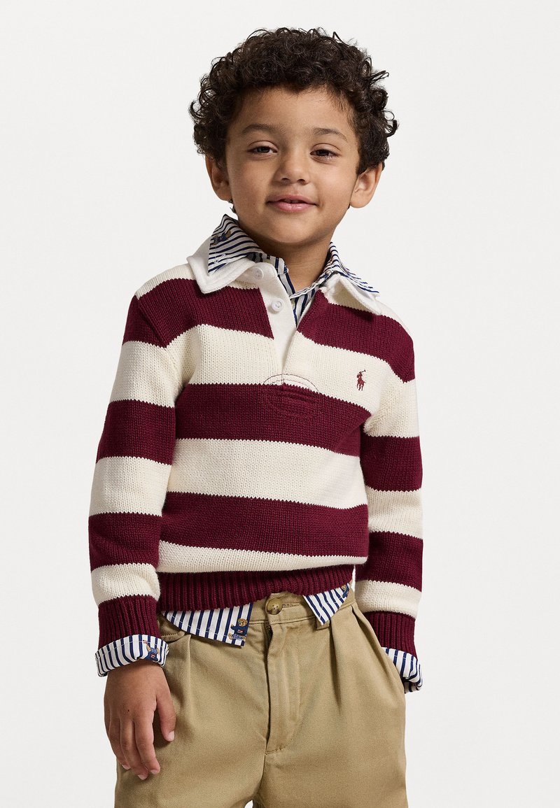 Ceriņsarkans un krēms svītrots adīts džemperis ar apkakles dekoltē, pārklāts ar zilu un balto svītraino krekli, kopā ar khaki biksēm.