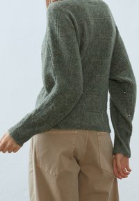 Grön stickad tröja med strukturerat mönster, långa ärmar och subtila pärlaccents. Matchad med beiga byxor, har en klassisk passform.