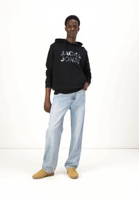 Felpa nera con cappuccio e logo "JACK & JONES", jeans blu e sneakers color sabbia. Vestibilità casual, tessuto testurizzato e taglio rilassato.
