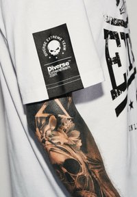 Biała koszulka z czarnym nadrukiem graficznym z napisem "Diverse Extreme Team". Posiada czarną metkę z nadrukiem czaski na rękawie. Złożony tatuaż widoczny na ramieniu.