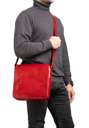 Homme portant un col roulé gris foncé et un jean noir, tenant un sac bandoulière en cuir rouge sur son épaule sur un fond blanc.