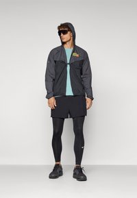 Vindjakke i sort med hætte, med logo-detalje, parret med en lyseblå skjorte, sorte shorts og sorte leggings. Sorte sko fuldender looket.