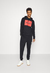 Svart hettegenser med rødt HUGO-logo, kombinert med svarte joggebukser med hvit tekst. Hvite sneakers med lyse tremøbler.