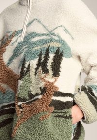 Polarowy pulower z teksturowanym, wielobarwnym wzorem krajobrazu z górami, drzewami i jeleniami. Obejmuje kołnierz na suwak oraz kieszenie na ręce.