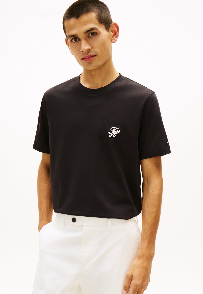 Νέος άνδρας που φοράει μαύρο κοντομάνικο μπλουζάκι με λευκό κεντημένο λογότυπο και λευκό παντελόνι, στέκεται μπροστά σε απλό φόντο.