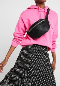 Persona che indossa una felpa rosa acceso, una borsa a tracolla nera con zip e una gonna nera con pois bianchi su uno sfondo semplice.