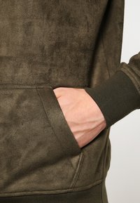 Hanorac din suede verde măsliniu, cu buzunar tip cangur, manșete ribbate și o textură fină, evidențiind materialul său moale și pufos.