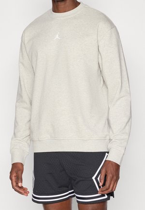 Mężczyzna w jasnoszarej bluzie z długim rękawem i okrągłym dekoltem z białym logo Jumpman oraz czarnych szortach do koszykówki z białymi paskami.