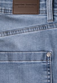Tejido de mezclilla azul claro, con un parche de marca de cuero, costuras dobles y un acento de botón plateado en el bolsillo delantero.