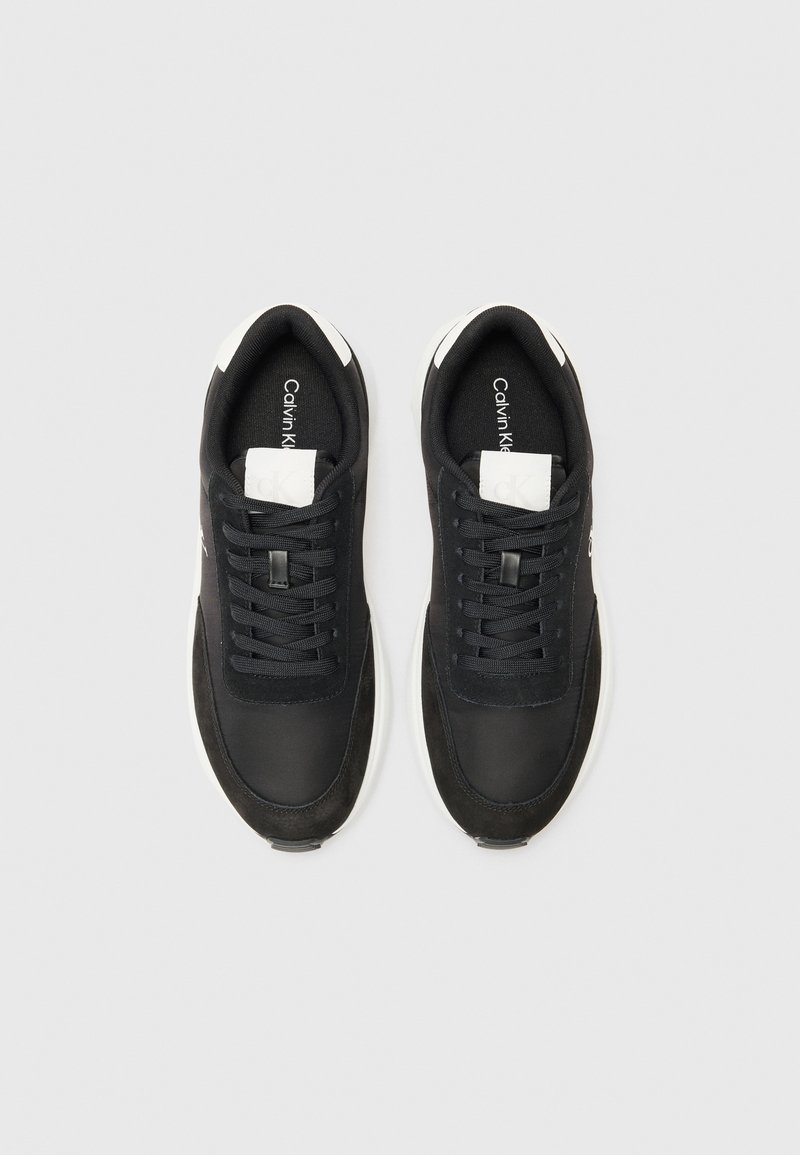 Baskets noires en mélange de matériaux lisses et texturés, avec une semelle blanche et un logo subtil sur la languette et le côté.