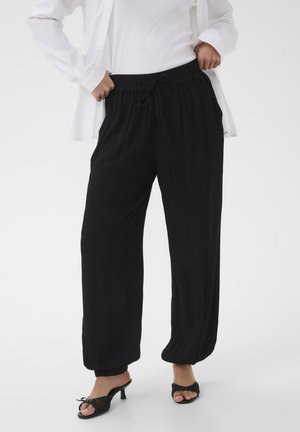 Femme portant un pantalon noir ample avec cordon de serrage et des sandales à talons noires, debout les mains dans les poches sur fond blanc.