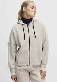 Šviesiai bežinė zip-up tipo palaidinė su priekinėmis kišenėmis, juodomis raištelėmis ir pintomis rankovėmis; derinama su juodu polo marškinėliu; laisvalaikio stilius.