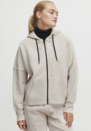 Lys beige zip-up hettegenser med frontlomme, sorte snorer og ribbestrikkede mansjetter; kombinert med svart genser med høy hals; uformell passform.