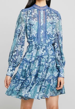 Femme portant une robe bleue à manches longues avec un motif, des détails en dentelle et une jupe à volants, debout devant un fond gris uni.