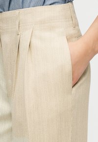 Pantalon beige texturé avec des plis et des passants de ceinture, porté avec une main dans la poche droite, associé à une chemise bleue.