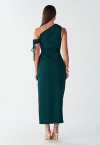 Abito da sera verde con una spalla sola, design arricciato, tessuto strutturato e laccetti sulla spalla. Abbinato a décolleté beige con cinturini sottili.