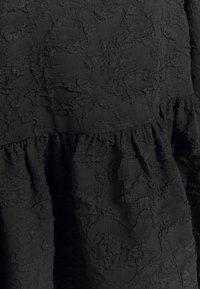 Svart klänning med texturerad tyg, som har ett skrynkligt mönster och lager av volangdetaljer längst ner. Mjukt, lätt material.