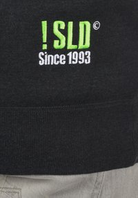Black sweatshirt featuring green and white embroidered text: “! SLD Since 1993.” Ribbed hem texture, simple design with no additional patterns.