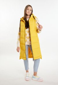 Gilet lungo imbottito giallo con cappuccio, chiusura a zip e cordini neri. Indossato sopra un maglione crema con motivi a cuore e jeans blu chiaro.
