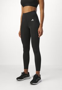 Czarne legginsy sportowe z wysokim pasem, z białym logo Adidas, subtelnymi detalami szycia i gładką, elastyczną teksturą materiału.