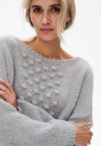 Jasnoszary sweter z dzianiny z teksturalnym wzorem bąbelków z przodu, szerokim dekoltem i oversize'owymi rękawami. Miękki, puszysty materiał.