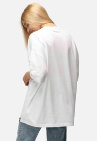 Bílý oversized bavlněný tričko s krátkými rukávy, kulatým výstřihem a detailem štítku na lemu. Nosené s světle modrými džíny.