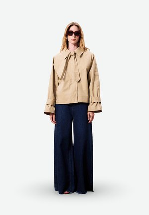 Femme portant une veste beige oversize avec un large col et un nœud, un pantalon large foncé, et de grandes lunettes de soleil foncées, debout droite sur un fond blanc.