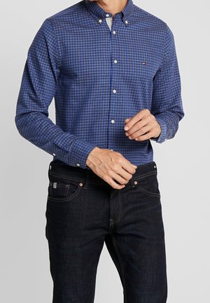 Muž v modrošedé kostkované košili na knoflíky a tmavých modrých džínách si upravuje manžety rukávů.