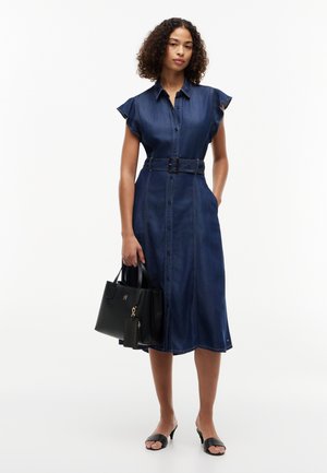 Donna in vestito midi di jeans con bottoni e cintura, che tiene una borsa nera, indossa décolleté nere aperte sulle punte su sfondo bianco.