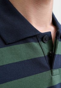 Roheline ja tumesinine rihmaga polo särk, mis sisaldab nööbiga kraed, tekstureeritud kangast ja sobivat disaini. Suurendatud vaade kaelaosa detailidele.
