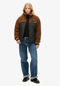 Veste en fausse fourrure brune et en tissu matelassé noir, avec une poche zippée, associée à un jean bleu et des chaussures noires. Design texturé avec des couleurs contrastantes.