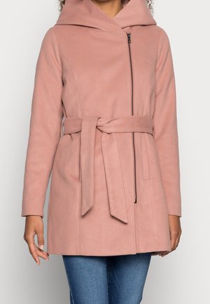 Kobieta w przybrudzonej różowej płaszczu z paskiem, z asymetrycznym przednim zamkiem błyskawicznym i kapturem, w połączeniu z niebieskimi dżinsami.