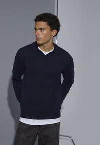 Marineblauer Strickpullover mit V-Ausschnitt und Kragen, kombiniert mit einem weißen Hemd darunter. Das Model steht mit Händen hinter dem Rücken vor einem neutralen Hintergrund.