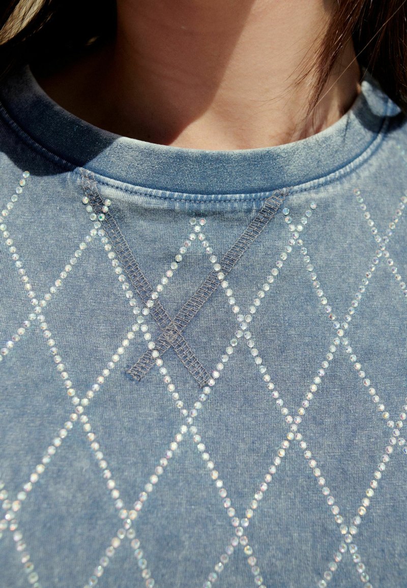Modrá denimová mikina se vzorem diamantů vyrobeným z iridescentních kamínků a kontrastními stehy kolem výstřihu.