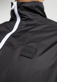 Must must jacket, mille kõrge krae ja valge tõmblukk. Märkimisväärsed detailid on tekstuuriga must ruudukujuline plaastriga, millel on "Urban Classics" logo.