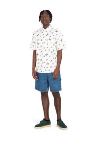 Camisa blanca de manga corta con patrones de mariposas de colores, combinada con pantalones cortos de mezclilla azules y zapatillas de ante verdes con suelas de crema.