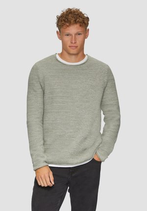 Lichtgroene gebreide trui met een gestructureerde afwerking, ronde halslijn en lange mouwen, gedragen over een wit shirt, gecombineerd met donkere jeans.